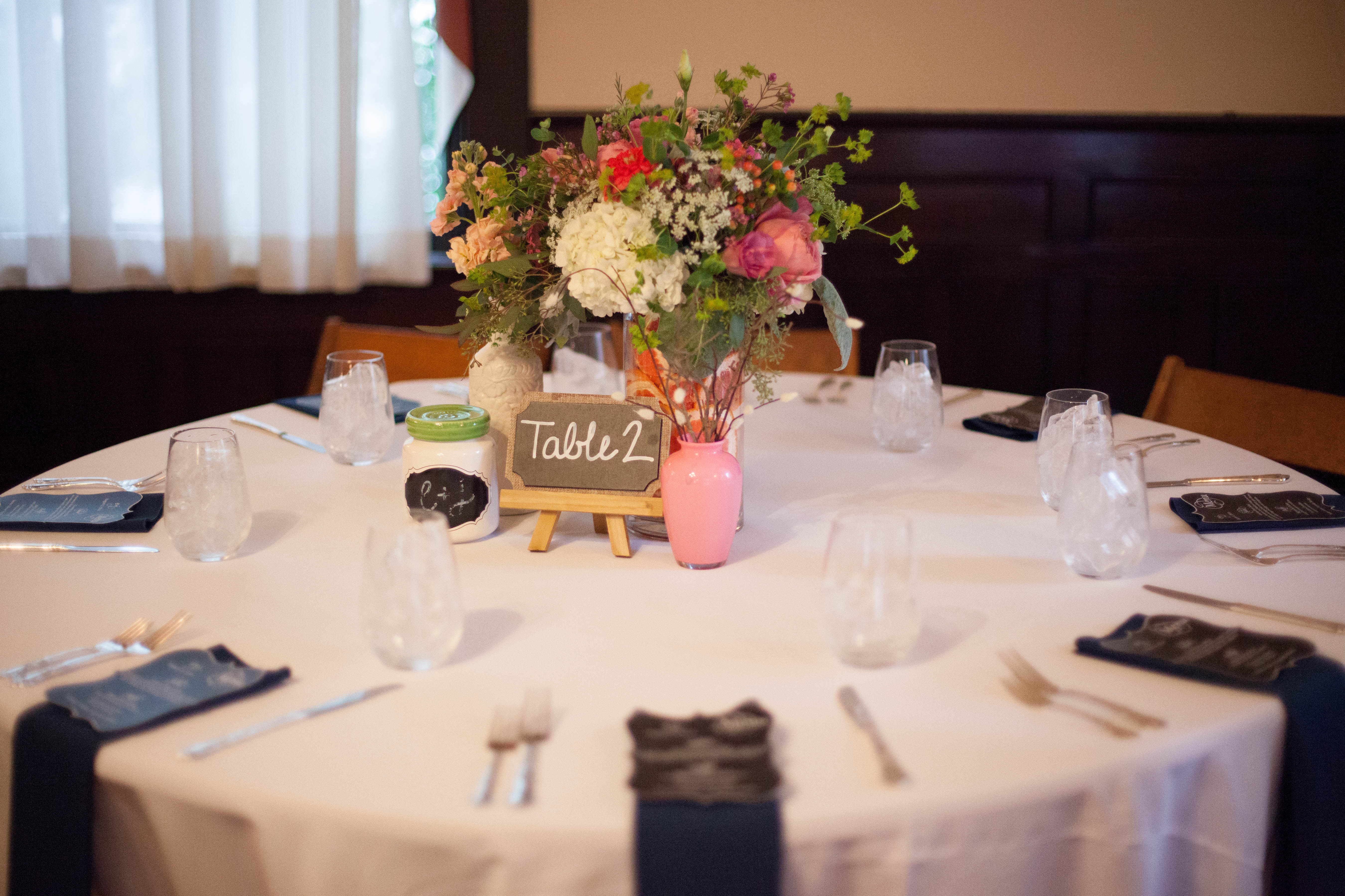 round guest table and white cotton linen with florals in painted bud vases from The English Garden Raleigh and table number on chalkboard with navy napkins and menu cards