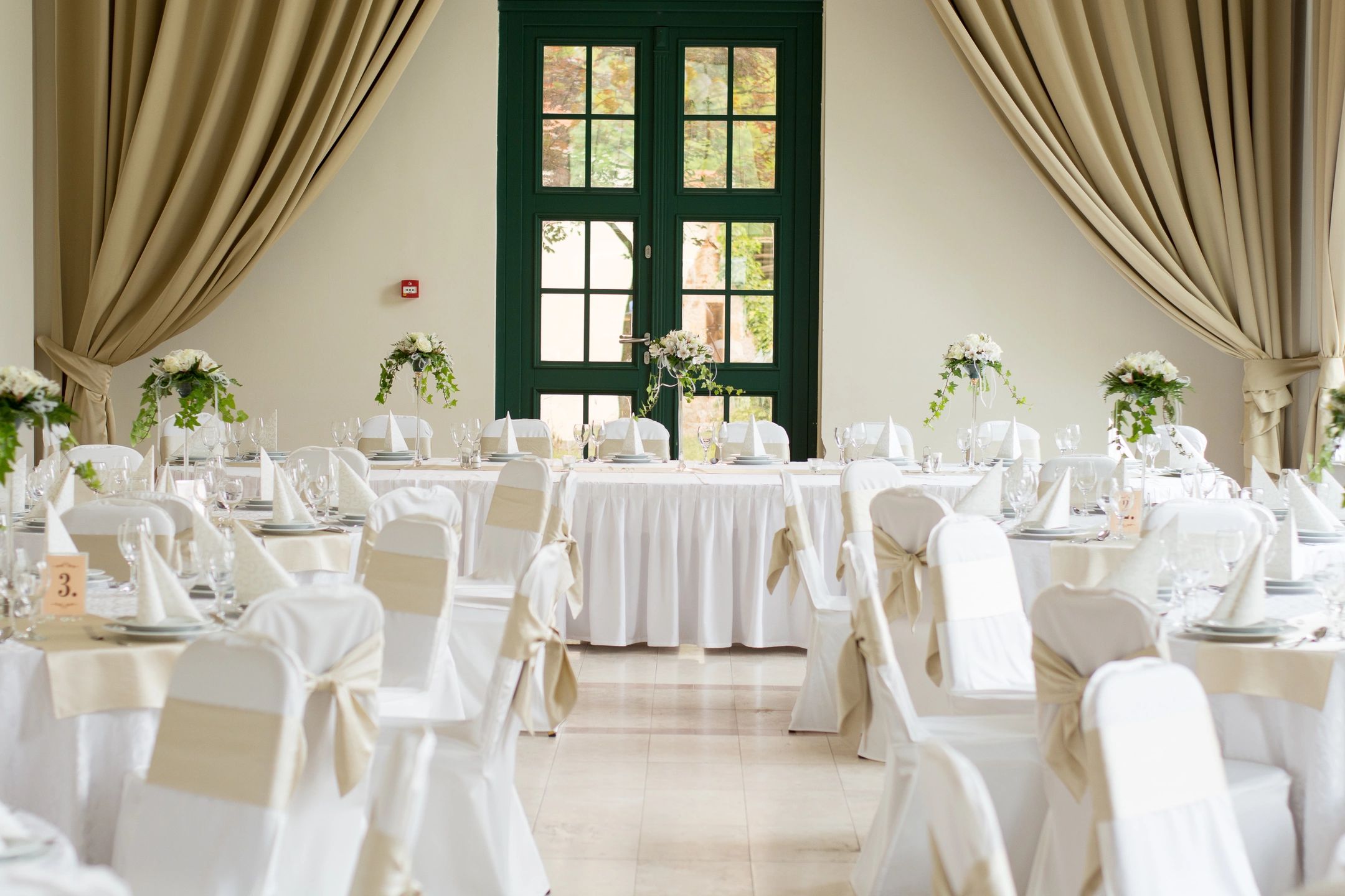 wedding reception room white and beige neutral with greenery centerpieces draping and green french doors